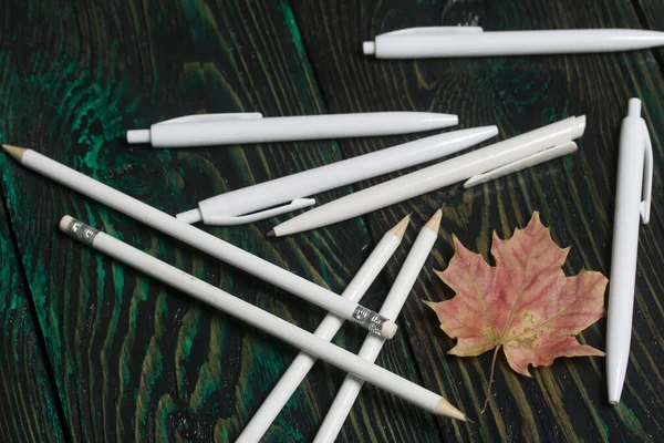 A set of white pencils and pens for school. They lie on pine boards. Nearby autumn maple leaves.