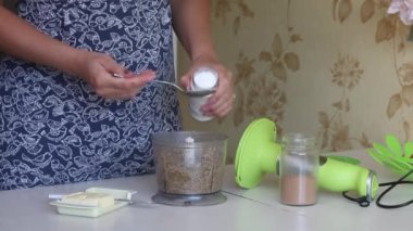 Bir kadın blender kasesinde doğranmış fındığa pudra şekeri ekler. Fındık ezmesi pişiriyorum. Ceviz kebabı. Malzemeler yan yana masada..