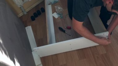 Adam mobilyaların parçalarını birbirine doğruluyor. Evde mobilya topluyor. Diğer parçalar yere saçılmış. Yukarıdan çekilen