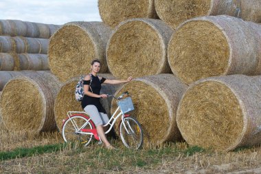 Bir saman yığınının yanında poz veren kız. Bisiklette oturuyor. Piramitte istiflenmiş saman ruloları.