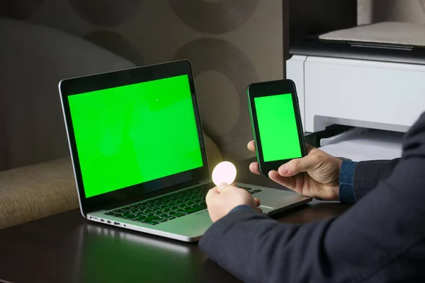 A man works at a laptop with a green screen. He holds a smartphone with a green screen and a cryptocurrency coin in his hand. Chromakey.