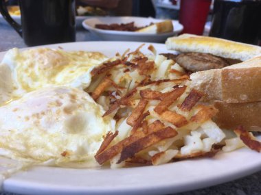 Bir lokantada kahvaltı. İki yumurta, kızarmış patates, kızarmış ekmek ve arka planda kahve fincanları olan beyaz bir tabakta sosisli börek. Tipik Amerikan kahvaltısı yol kenarındaki bir lokantada.
