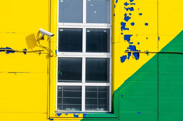 A video camera next to a white-framed window on a facade painted with peeling yellow, green, and blue paint.