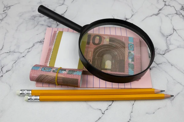 Ofis malzemeleriyle dolu 10 avroluk banknotların üzerinde bir büyüteç duruyor..
