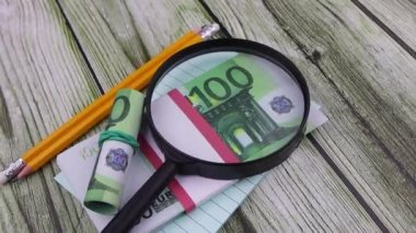 Ofis malzemeleriyle dolu 100 avroluk banknotların üzerinde bir büyüteç duruyor..
