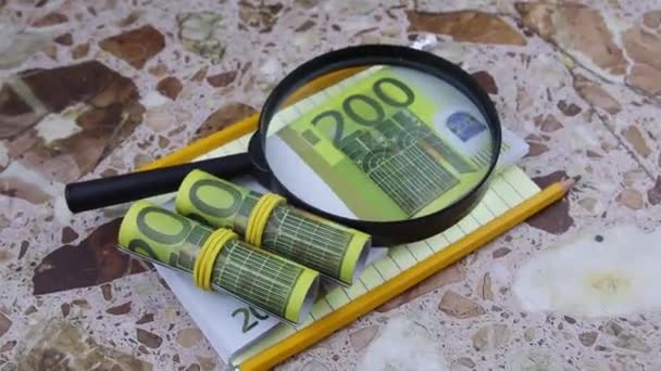 Ett förstoringsglas ligger på ett paket med 200 eurosedlar med kontorsutrustning. — Stockvideo