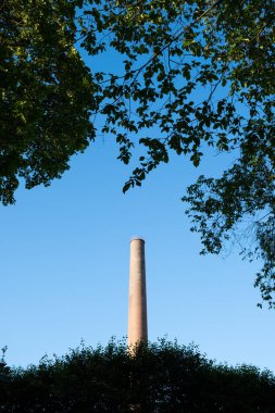 Eski sanayi, mavi gökyüzüne karşı yaprakların arasından görünen baca.