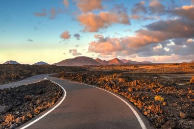 Lanzarote, Kanarya Adaları 'nda açık yol... Tepeleri, dağları ve volkanik kraterleri olan kayalık bir çölde... Pembe bulutlarla dolu mavi bir gökyüzünün altında....