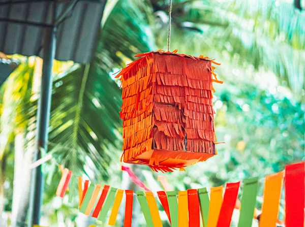 Quadratisch geformte kinderparty mexikanische orange pinata hängen draußen für spaß in posadas und geburtstagsferien, bunte gelb-rot-grüne dekorationsfahnen verwendet. Natur Palmen Garten Hintergrund Stockbild