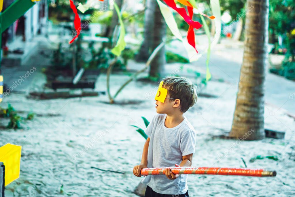 Side view little kid boy wearing lego style yellow face mask gesturing preparing to hit pinata with big long stick outdoor on birthday party. Nature palm trees garden background, red decoration flags