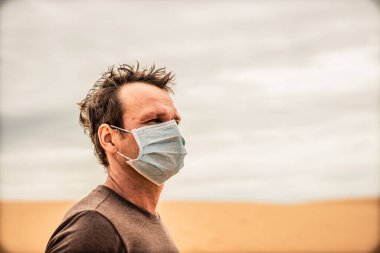Yan portre genç adam koruyucu maske takıyor, yüz ifadesi duygu uzak kum tepeciği gibi görünüyor. Konsept COVID stres sendromu, akıl sağlığı, tehlike kirliliği korkusu, olumsuz sosyo ekonomik sonuç