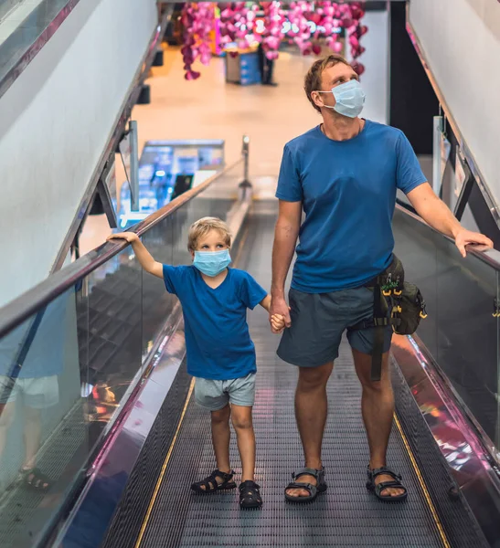 Pemandangan depan anak laki-laki, ayah naik eskalator di super market memakai wajah melindungi topeng, COVID coronavirus pandemic karantina kuncian. Ayah memegang tangan anak, melihat ke atas. Realitas kehidupan baru, belanja keluarga — Stok Foto