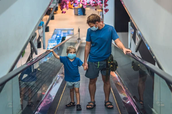 Front view pojke, far går uppför rulltrappan i snabbköpet slitage ansikte skydda mask, COVID coronavirus pandemisk låsning karantän. Pappa håller barnets hand, titta på ungen. Nytt liv verklighet, familj shopping Stockbild