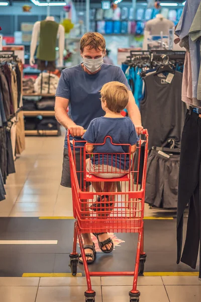 Ayah serius menggulung anak laki-laki di troli gerobak toko merah, memakai topeng wajah biru melindungi dari virus COVID, ukuran kontrol kesehatan pribadi publik, belanja keluarga selama pandemi, membeli pakaian baru — Stok Foto
