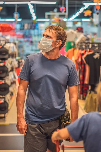 Ayah serius menggulung anak laki-laki di troli gerobak toko merah, memakai topeng wajah biru melindungi dari virus COVID, ukuran kontrol kesehatan pribadi publik, belanja keluarga selama pandemi, membeli pakaian baru — Stok Foto