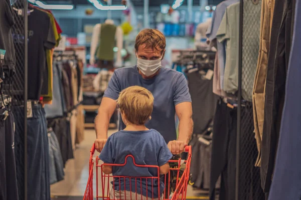 Серьезный отец катит ребенка мальчик в красном магазине тележку, носить синюю маску для лица защиты от вируса COVID, общественный личный контроль здоровья, семейные покупки во время пандемии, покупка новой одежды Стоковое Фото