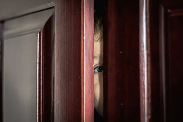 Sorglig pojke gömma sig i garderoben, närbild av blått öga i en glänsande dörr slit hem garderob. Barnkänslor. Varningstecken, behandling av depression hos barn, rädsla, familjelivet Stockbild