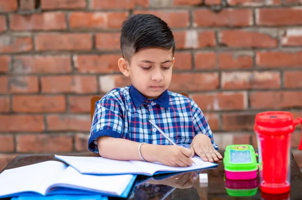 Cute little Indian/Asian school kid studying on study table , writing on notebook with pencil.