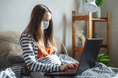 Koruyucu maskeli genç bir kız evde bir yatakta oturuyor ve bir dizüstü bilgisayar kullanıyor. Uzaktan öğrenme, uzaktan çalışma, salgında online iletişim kavramı.