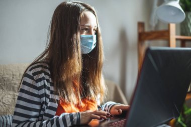Koruyucu maskeli genç bir kız evde bir yatakta oturuyor ve bir dizüstü bilgisayar kullanıyor. Uzaktan öğrenme, uzaktan çalışma, salgında online iletişim kavramı.