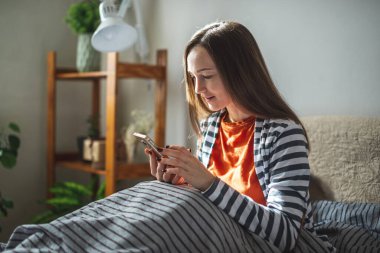 Genç kız gün ışığında yatağında oturuyor ve iletişim kurmak ya da bilgi aramak için telefonunu kullanıyor. Günün başlangıcı ve yaşam tarzı kavramı.