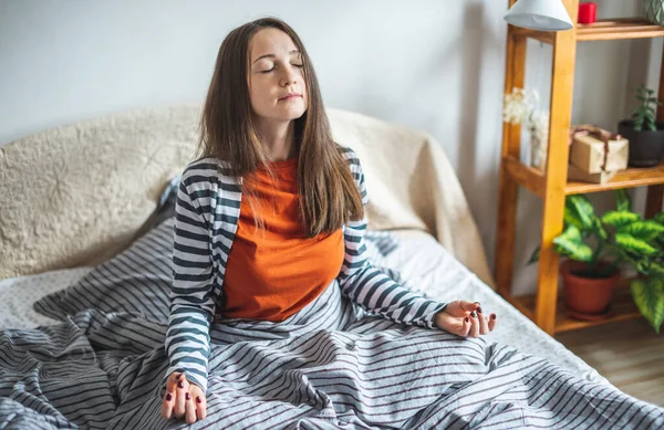 A young woman in pajamas is sitting on the bed in the morning in the lotus position and meditating. Concept of mood for a good day.