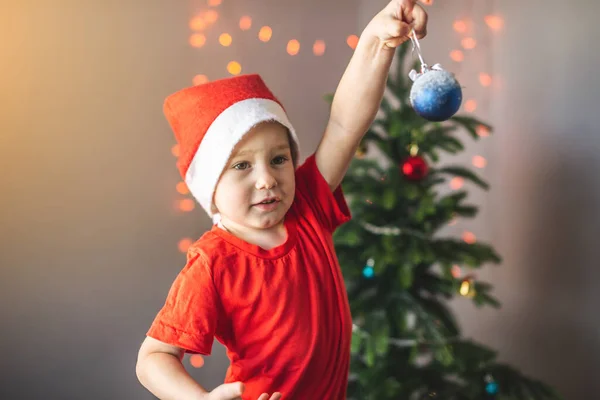 サンタの帽子のかわいい子供は ガーランドが飾られたクリスマスツリーの背景にクリスマスボールを手に持っています — ストック写真