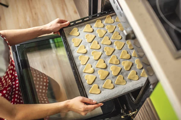 Woman is putting a baking sheet with delicious homemade cookies in the shape of a heart in the oven. Concept of fresh homemade cakes or a surprise for Valentine\'s Day.