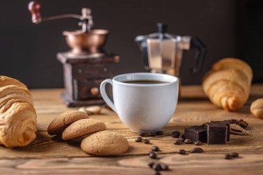 Tahta bir masada kruvasan, kurabiye ve çikolatalı sade kahve var. Arka planda bir kahve değirmeni ve gayzer kahve makinesi var..