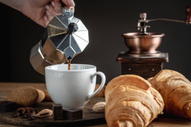 Bir el gayzer kahve makinesinden beyaz bir fincana siyah aromatik kahve döküyor. Taze kruvasan ve kahve öğütücüsü ahşap masadaki fincanın yanında..