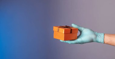 Mavi koruyucu eldivenli bir adam mavi ve gri arka planda bir hediye kutusu tutuyor. Bir salgında yaşam tarzı kavramı.