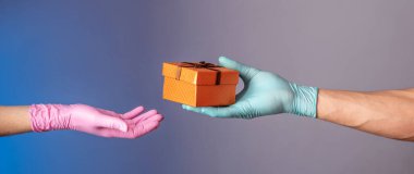 Mavi eldivenli bir adam mavi ve gri arka planda pembe eldivenli bir kadının eline hediye kutusu veriyor. Bir salgında yaşam tarzı kavramı.