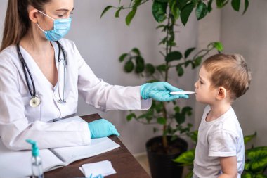Genç bir bayan doktor, küçük bir çocuğun ateşini elektronik termometre kullanarak ölçüyor..