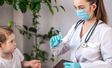 Beyaz elbiseli, mavi eldivenli ve koruyucu maskeli genç bir bayan doktor küçük bir erkek hastaya aşı yapıyor. Sağlık ve aşı kavramı.