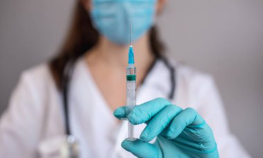 Beyaz elbiseli, mavi eldivenli, koruyucu maskeli ve steteskoplu genç bir bayan doktor elinde aşı olan bir şırınga tutuyor. Sağlık hizmeti ve aşı kavramı. Yakın çekim..