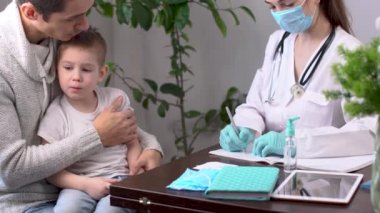 Babasıyla birlikte küçük bir çocuk çocuk doktorunu ziyaret ediyor. Bir doktor hasta bir çocuk için reçete yazıyor.