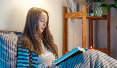 Genç bir kadın yatmadan önce yatakta bir kitap okuyor. Uyku zamanı kavramı, dinlenme ve sağlıklı uyku için uyku ritüelleri..