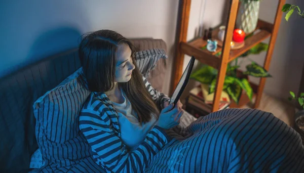 A young woman in a dark bedroom is laying in bed late at night and using her tablet to surf the Internet or read an electronic book. Concept of bedtime pastime and insomnia.