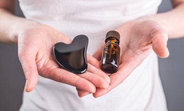 Bir kadın elinde masaj aleti, doğal taştan yapılmış bir guaj kazıyıcı ve yüz masajı ve cilt bakımı için bir şişe organik aromatik yağ tutuyor. Yakın çekim..