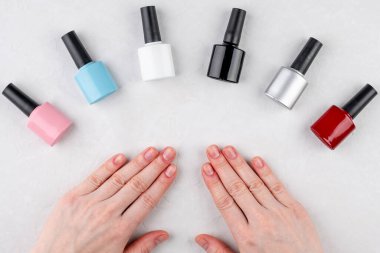 Farklı renklerde oje şişeleri ve beyaz arka planda montsuz kısa tırnaklı eller. Manikür ve renk seçimi kavramı. Üst görünüm.