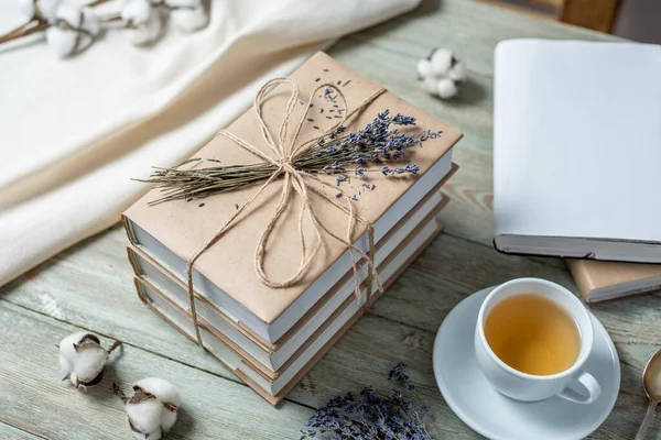 Cute stack of books with craft paper covers, wrapped in rope and decorated with lavender flowers, and a cup of tea on a wooden table. Concept of a cozy atmosphere for reading.