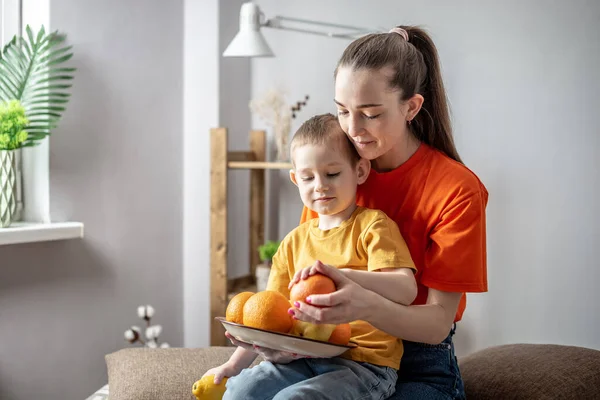 Młoda Mama Małe Dziecko Jasnych Ubraniach Siedzą Talerzem Pomarańczy Cytryn — Zdjęcie stockowe