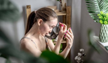 Elinde ayna tutan ve yüzünde kozmetik siyah bir maskeyi çıkarmakta zorlanan bir kadının komik fotoğrafı. Cilt bakımı kavramı, evdeki kozmetik prosedürleri, yaşam tarzı.