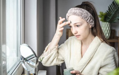 Bornozlu genç ve güzel bir kadın yüzüne yeşil bir kozmetik maske takıyor. Cilt bakımı ve yaşam tarzı kavramı.