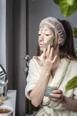 Bornozlu genç ve güzel bir kadın yüzüne yeşil bir kozmetik maske takıyor. Cilt bakımı ve yaşam tarzı kavramı.