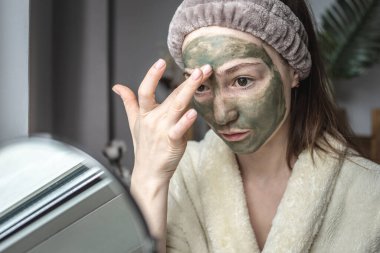 Aynanın önünde bornozlu genç ve güzel bir kadın yüzüne yeşil bir kozmetik maske takıyor. Cilt bakımı kavramı, doğal kozmetik kullanımı ve yaşam tarzı.