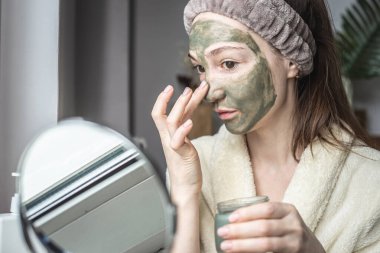 Aynanın önünde bornozlu genç ve güzel bir kadın yüzüne yeşil bir kozmetik maske takıyor. Cilt bakımı kavramı, doğal kozmetik kullanımı ve yaşam tarzı.