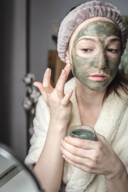 Bornozlu genç ve güzel bir kadın yüzüne yeşil bir kozmetik maske takıyor. Cilt bakımı ve yaşam tarzı kavramı.