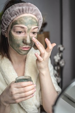 Aynanın önünde bornozlu genç ve güzel bir kadın yüzüne yeşil bir kozmetik maske takıyor. Cilt bakımı kavramı, doğal kozmetik kullanımı ve yaşam tarzı.