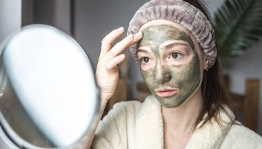 Bornozlu genç ve güzel bir kadın yüzüne yeşil bir kozmetik maske takıyor. Cilt bakımı ve yaşam tarzı kavramı.